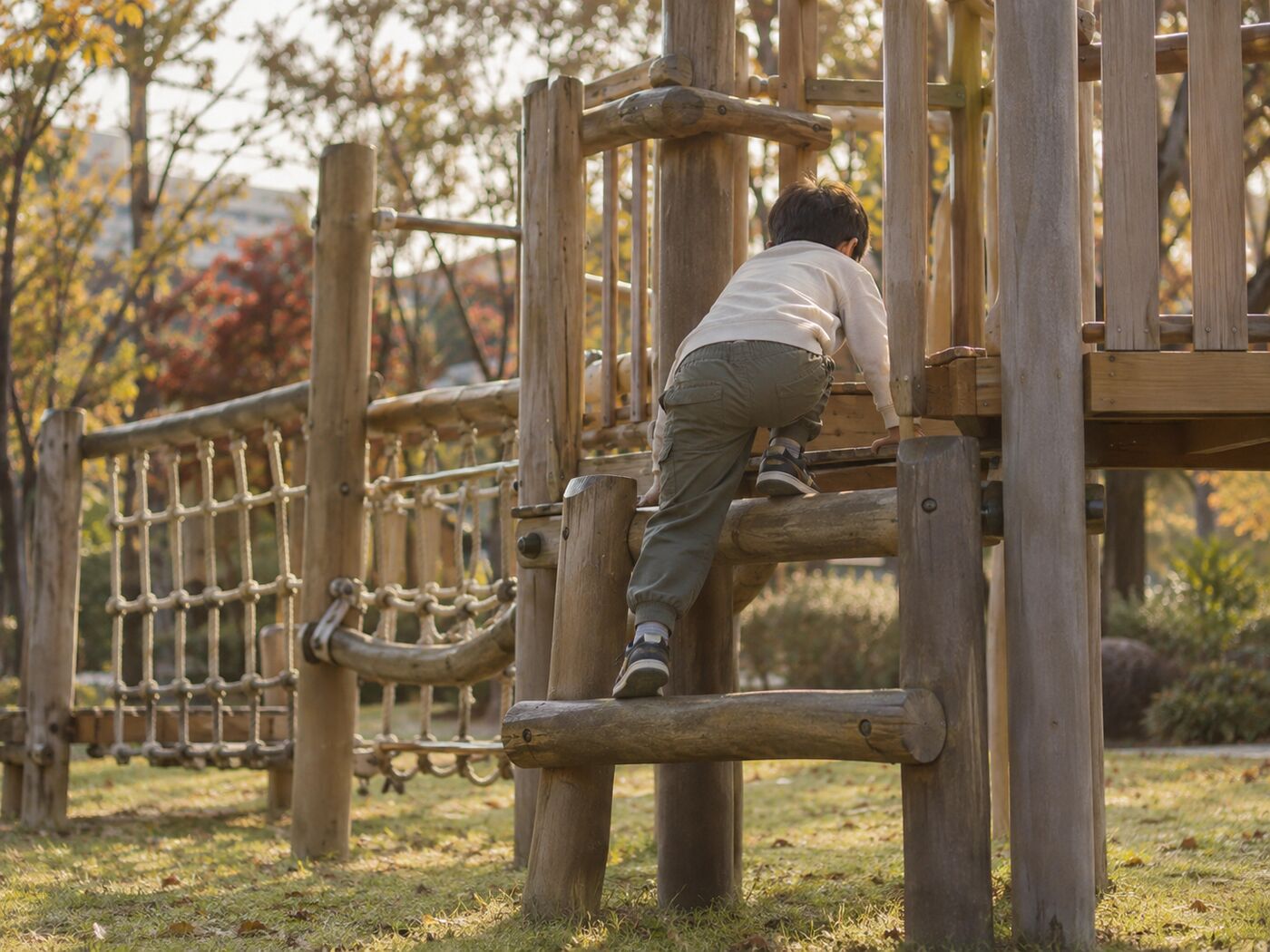 공원 잔디밭에 설치된 나무색 야외 놀이 구조물을 오르는 여덟 살쯤 되는 남자아이의 뒷모습